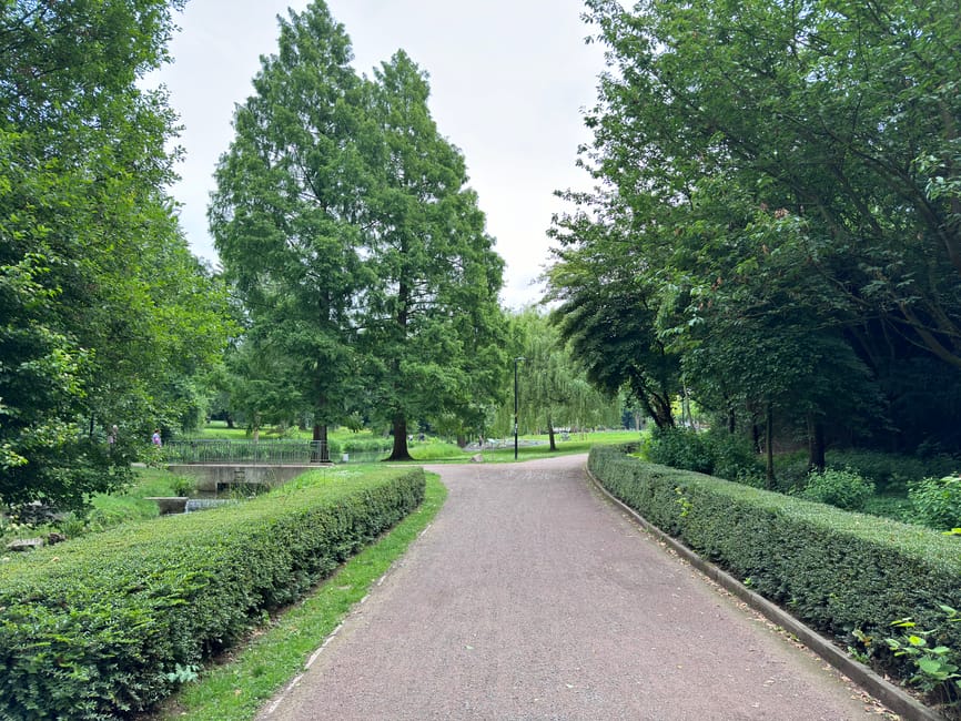 A paved pathway winding through a well-maintained garden area, bordered by neatly trimmed hedges and lush green trees. The pathway appears to be composed of fine gravel or asphalt, with a smooth surfa