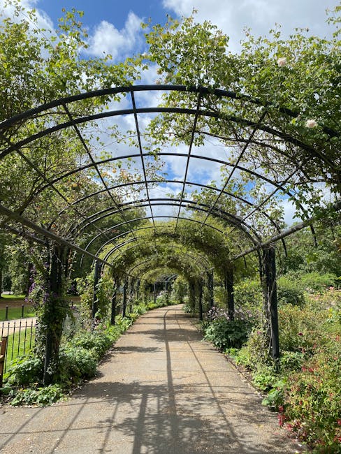 A paved pathway winding through a well-maintained garden area, bordered by neatly trimmed hedges and lush green trees. The pathway appears to be composed of fine gravel or asphalt, with a smooth surfa